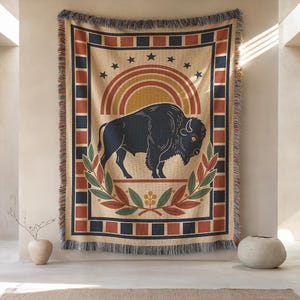 May include: A woven throw blanket with a buffalo design. The blanket features a tan background with a navy blue buffalo, a rainbow, and stars. The border has a geometric pattern in red, navy, and tan. The blanket has a fringed edge.
