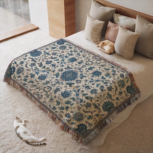 May include: A woven throw blanket with a floral pattern in shades of blue and cream, fringed edges, and a decorative design. The blanket is draped over a bed with pillows. A cat is lying on the bed and another cat is lying on the floor.