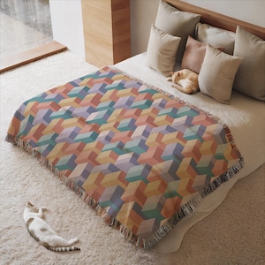 May include: A colorful geometric patterned throw blanket in shades of peach, teal, yellow, and purple, with a fringed edge. The blanket is draped over a bed with pillows and a sleeping cat. A second cat is lying on the carpet.