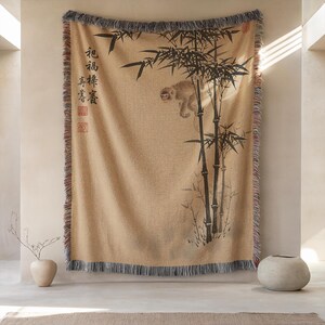 May include: A woven throw blanket in a tan hue featuring a black ink-style illustration of a monkey climbing bamboo stalks. The design includes vertical Asian calligraphy characters on the left side and a fringed edge with multicolored threads.