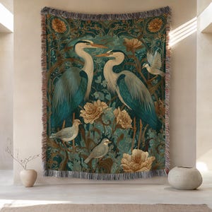 May include: A woven tapestry featuring a teal background with a design of herons and birds amidst floral elements. The herons are depicted in shades of blue and white, with tan flowers and a fringed border.
