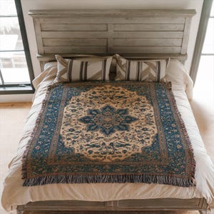 May include: A blue and beige patterned throw blanket with fringe, draped over a bed. The blanket features a central floral design and a border with intricate patterns. Two striped pillows rest against the headboard.