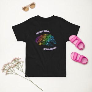 May include: Black toddler t-shirt featuring a rainbow spaceship graphic and the words "Cutest Rebel in the Galaxy." The image also shows pink sandals and sunglasses.