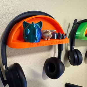 Puede incluir: Dos soportes para auriculares impresos en 3D con estantes. Los estantes son de color naranja y verde y tienen el texto "Mr. Ginger's Workshop" impreso en ellos. Los soportes están montados en una pared y tienen auriculares negros colgando de ellos.