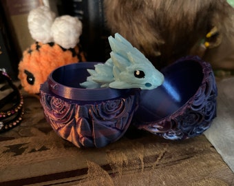 Può includere: Una scatola per gioielli a forma di uovo blu e viola con un motivo di rose dettagliato. La scatola è aperta e all'interno c'è una piccola statuetta di drago blu.