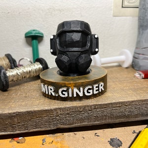 May include: A black 3D printed figurine of a head wearing a gas mask, sitting on a wooden base with the words "MR. GINGER" written on it.