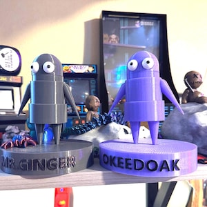 May include: Two 3D printed figurines, one gray and one purple, stand on bases with the names "Mr. Ginger" and "OkeeDoak" printed on them.
