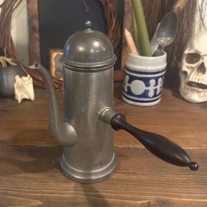 Könnte beinhalten: Eine Kaffeekanne aus Zinn mit einem schwarzen Holzgriff. Die Kanne hat einen abgerundeten Deckel und einen Ausguss, der sich nach oben krümmt. Die Kanne steht auf einer Holzoberfläche.
