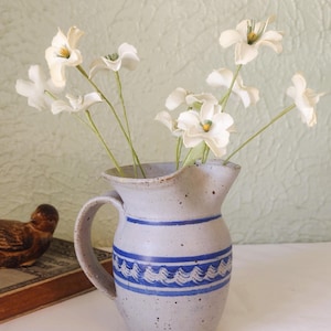 May include: A speckled light gray ceramic pitcher with a blue horizontal band featuring a white wave pattern. The pitcher has a curved handle and a pouring spout, and it contains several white artificial flowers with yellow centers.