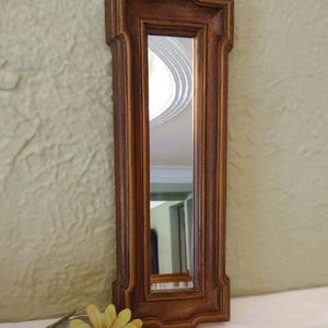 May include: A rectangular wall mirror featuring a dark brown wooden frame with decorative carved edges and a vertical orientation. The mirror surface reflects a bright interior space, and a single artificial yellow flower rests nearby.