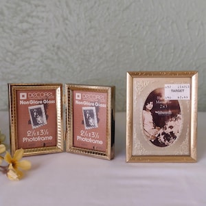 May include: Three gold-colored photo frames. One is a double frame, hinged in the middle, with the text "DECOREL Non Glare Glass" and "2 1/2 x 3 1/2 Photoframe". The other is an oval frame with a photo and a price tag.