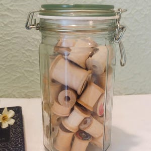 May include: A glass jar filled with wooden cotton spools. The jar has a green lid and metal clasp. A small, antique book with a flower is visible to the left. The spools are various sizes and shapes.