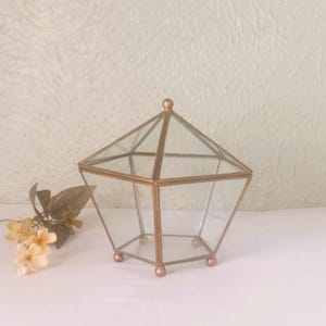 May include: A geometric terrarium with a copper-colored metal frame and clear glass panels. The terrarium has a pentagonal shape with a small, round finial on top and sits on small, round feet. A few dried flowers are to the left.