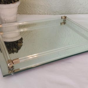 May include: Rectangular mirrored tray with clear acrylic and gold-tone metal accents. The tray has a clear acrylic frame with gold-tone metal corner details. The mirrored surface reflects the surrounding objects. A decorative item is in the background.
