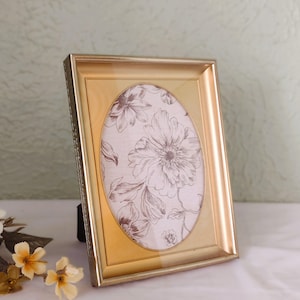 May include: A gold-colored, rectangular picture frame with an oval opening. The frame holds a floral print in shades of brown and cream. The frame is propped up on a black stand, and sits on a white surface.