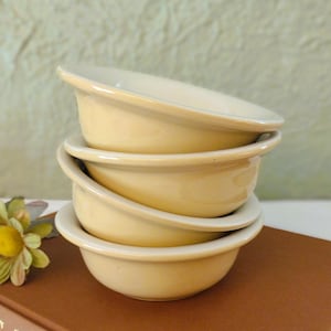 May include: A stack of four small, round, cream-colored ceramic bowls with smooth, curved edges. The bowls are uniform in size and shape, featuring a glossy finish that reflects light. They are stacked vertically, creating a consistent profile.
