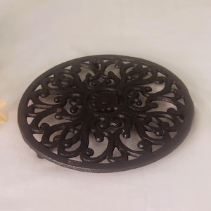 May include: A dark brown cast iron trivet with an intricate floral design. The round trivet has a raised edge and small feet. This decorative piece is designed to protect surfaces from hot cookware.
