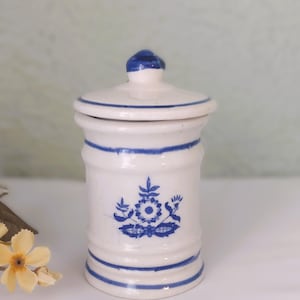 May include: A small, white ceramic jar with a lid, adorned with blue floral designs and horizontal blue stripes. The jar has a cylindrical shape and a small knob on top of the lid. The overall aesthetic is vintage and decorative.