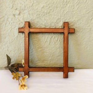 May include: A square wooden frame with a dark brown finish and extended corner joints. The frame features a grid-like construction with four intersecting bars. A small sprig of dried yellow flowers is positioned near the bottom left corner.