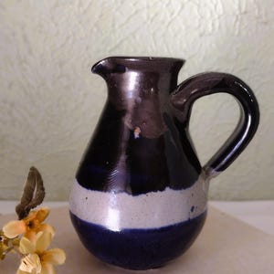 Peut inclure: Pichet en céramique fait à la main, avec un dessus marron foncé, une bande blanche et une base bleu foncé. Le pichet a une anse incurvée et un bec verseur. Des fleurs décoratives sont au premier plan.