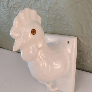 Peut inclure: Un crochet mural en céramique blanche en forme de tête de coq. Le coq présente une finition brillante, une crête proéminente et un œil orange visible. Un petit crochet métallique dépasse de la base rectangulaire dotée d'un trou de fixation.