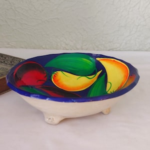 May include: A small white ceramic bowl with three feet, hand-painted with a blue rim and colourful peppers and leaves.