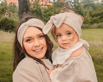 May include: A woman wearing a beige jacket and a beige headband holds a young child wearing a beige sweater and a beige headband. The child is wearing a white bib and floral leggings.