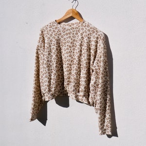 Peut inclure: Un pull à manches longues de couleur crème avec un motif léopard brun. Le pull est suspendu à un cintre en bois sur un mur blanc.