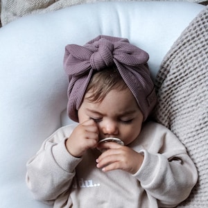 Könnte beinhalten: Ein Baby mit einem mauvefarbenen Turban mit Schleife, einem beigen Sweatshirt mit dem Text "du.." und einer passenden Hose. Das Baby hält einen Schnuller und liegt auf einem weißen Kissen und einer strukturierten Decke.