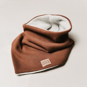 May include: A brown waffle knit neck warmer with a white fleece lining. The warmer has a brown leather label with the text "Stuck Glück" on it.