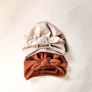 Op de afbeelding: Twee stoffen hoofdbanden met een geknoopt strikdesign. De bovenste hoofdband is licht beige van kleur en de onderste hoofdband is oranje-bruin.