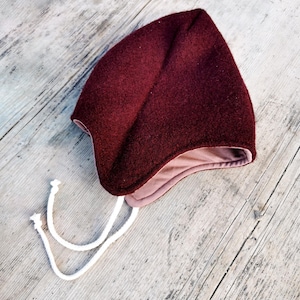 May include: A burgundy baby bonnet with a soft, textured exterior and a light pink interior. The bonnet has a pointed top and ear flaps, with white drawstrings for securing. The hat is made of a thick, warm material.