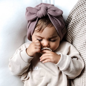 Könnte beinhalten: Ein Baby trägt einen mauvefarbenen Turban mit einer großen Schleife oben und einen beigefarbenen Pullover mit dem Wort "du." darauf gedruckt. Das Baby hält einen weißen Schnuller.
