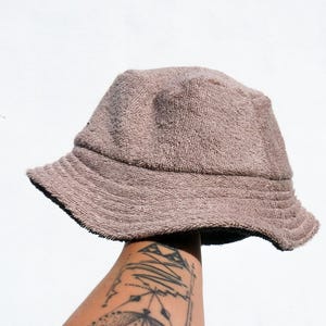 Może przedstawiać: Brązowy kapelusz typu bucket wykonany z frotte. Kapelusz jest trzymany przez rękę z geometrycznym tatuażem.