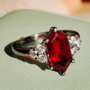 Op de afbeelding: Een zilveren ring met een grote, facetgeslepen, rode edelsteen in een zeshoekige vorm, geflankeerd door kleinere, ronde, heldere stenen. De ring ligt op een lichtgroen oppervlak, met zachte verlichting.