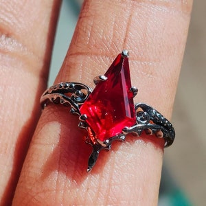 May include: A silver ring with a large, red, faceted gemstone in the shape of a point. The ring has a delicate, ornate design.