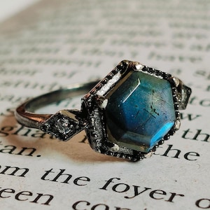 May include: A silver ring with a hexagonal, iridescent blue-green gemstone. The ring has a detailed, antique-style setting with small accent stones. The ring is set against a backdrop of a book page.
