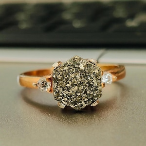 Op de afbeelding: Een gouden ring met een zeshoekige pyriet edelsteen en twee kleine heldere stenen.