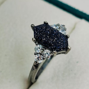 Peut inclure: Une bague noire avec une pierre hexagonale bleu foncé scintillante et de plus petites pierres claires autour.