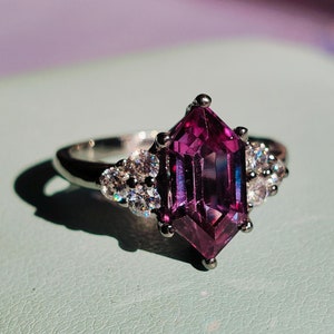 Op de afbeelding: Een zilveren ring met een grote, gefacetteerde, paarse edelsteen omringd door kleinere, heldere edelstenen.