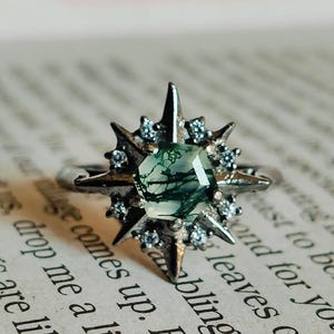 May include: A silver ring with a green moss agate stone set in a starburst design. The ring is on a book with text.