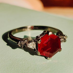 May include: A silver ring with a large, red, hexagonal gemstone surrounded by smaller, clear gemstones.