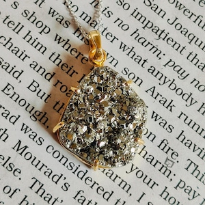 Puede incluir: Un collar de plata con un colgante con una piedra brillante de color plateado, de forma irregular. El colgante está enmarcado con metal dorado y cuelga de una delicada cadena de plata. El fondo es una página de texto impreso.