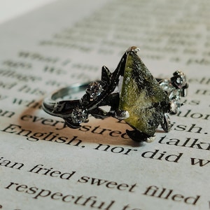 Pode incluir: Um anel de prata com uma grande gema verde facetada engastada em uma moldura de metal preto. O anel é adornado com detalhes florais delicados e pedras preciosas menores.