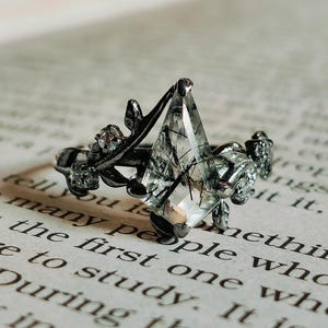 May include: A silver ring with a clear, faceted gemstone set in a black metal setting. The ring is adorned with delicate black metal floral accents.