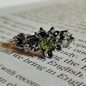May include: A silver ring with a green gemstone set in a flower design. The ring is on a book with text.