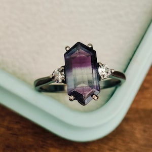 May include: A silver ring featuring a large, faceted, purple and green gemstone. The gemstone is surrounded by two smaller, clear gemstones.