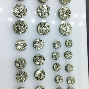 Peut inclure: Une collection de 20 cabochons ronds et plats en pyrite de couleur argentée, disposés en trois rangées sur une surface blanche. Les cabochons sont de tailles variées et présentent une texture cristalline scintillante.