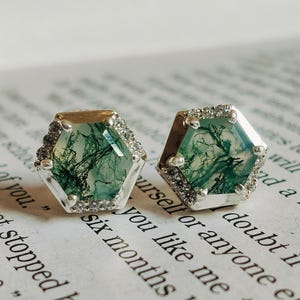May include: A pair of silver earrings with hexagonal green moss agate stones set in a silver bezel. The stones have a unique, natural pattern of green and white.
