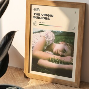 May include: A framed poster with a minimalist design featuring a photo of a young woman lying in a field of grass. The poster is titled "The Virgin Suicides" and includes the year 1999 and the runtime of 97 minutes.